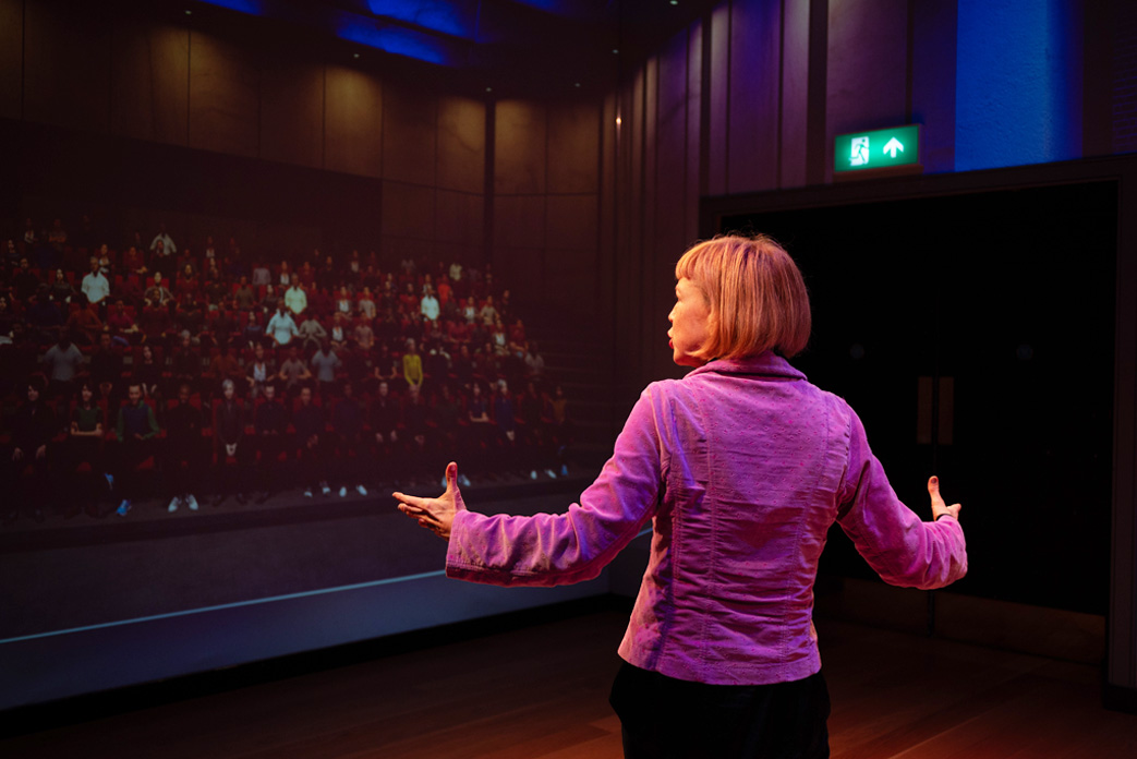 A women wearing a purple blazer, speaking to a virtual audience simulated on a wall.